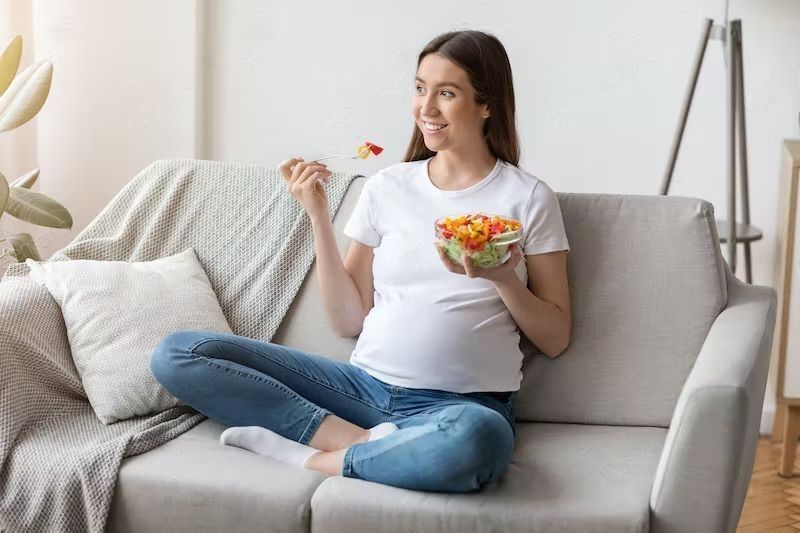 What To Eat When Pregnant? A young expectant woman enjoying fresh vegetable salad while relaxing on couch at home