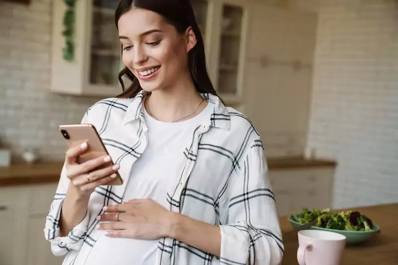 Understanding The Ins And Outs Of Pregnancy. A happy pregnant woman using cellphone while having breakfast in cozy kitchen