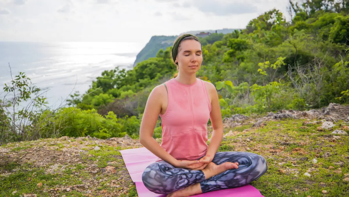 Young woman sitting on yoga mat in lotus pose, meditating, practicing yoga and pranayama. Deep breath and concentration. Outdoor yoga on the cliff. Bali, Indonesia