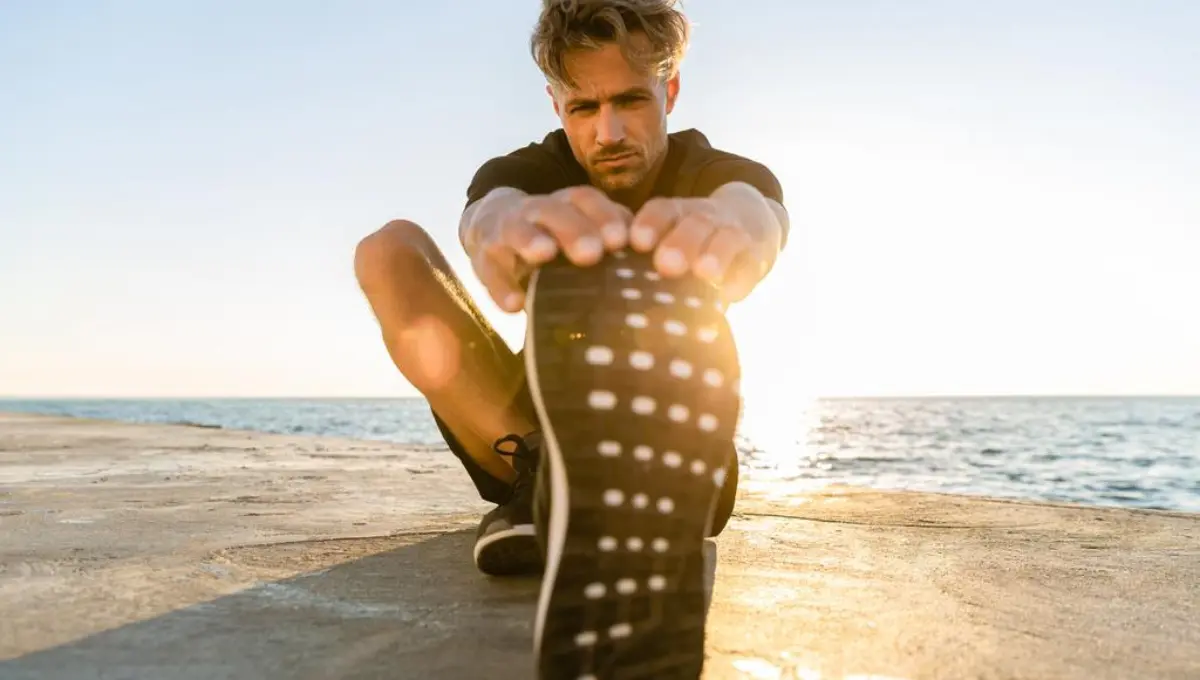 Sporty adult man stretching leg on seashore in front of sunrise