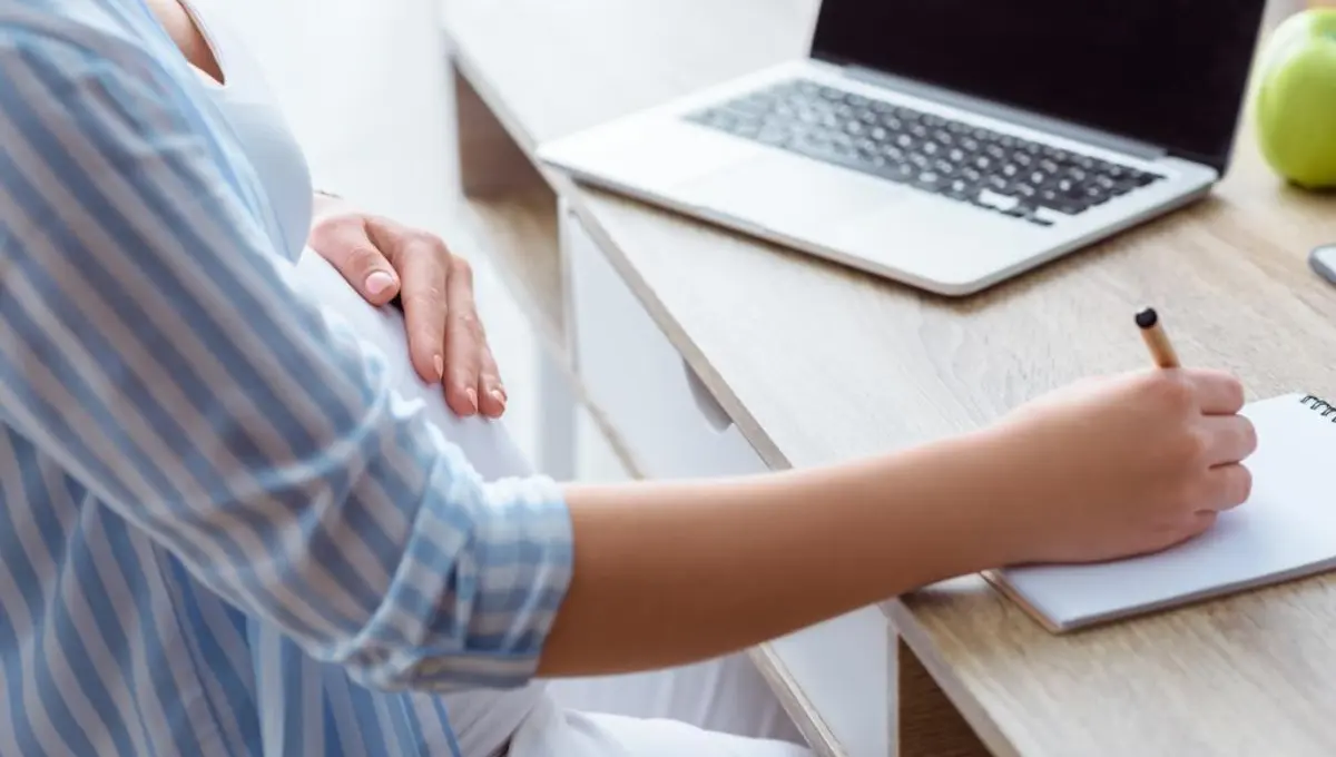 a pregnant woman writing in notepad and using laptop at home