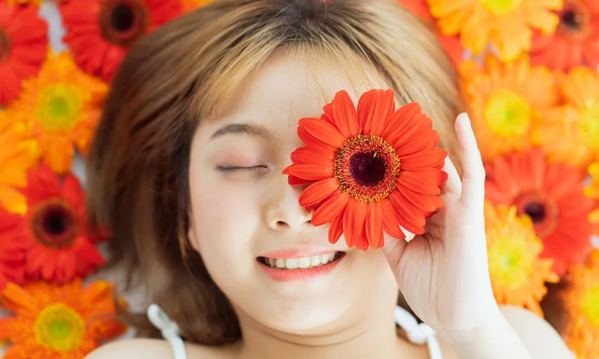 a woman lying on flowers with 1 orange colored marigold flower in her hand above her eye.